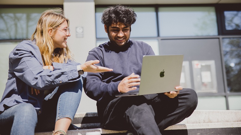 Two Oxford Brookes University students sitting outside on campus, smiling as they look at a laptop together during a collaborative study session.