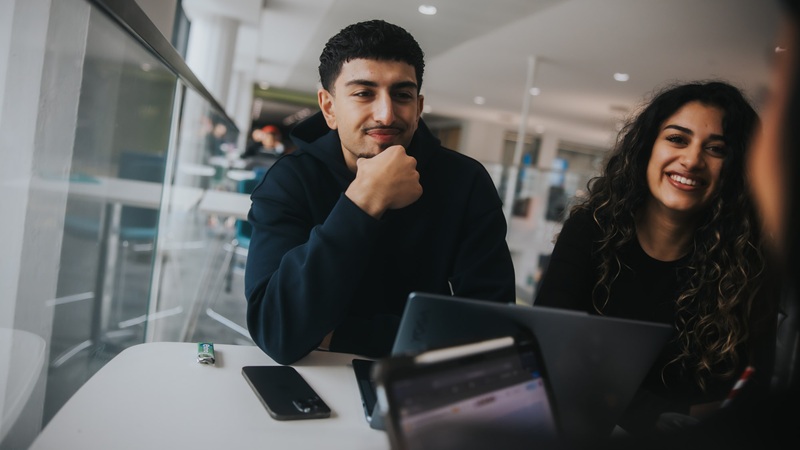 Two students smiling and talking with their laptops on a desk