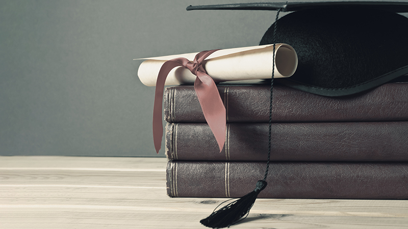 A rolled diploma tied with a ribbon resting on a stack of vintage books next to a black graduation cap.