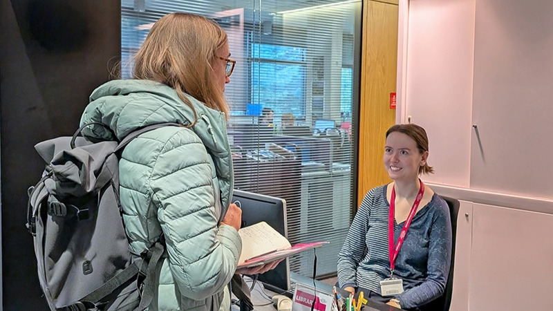 A library colleague sits behind a computer at a desk. A library user stands in front of her, holding an open notebook