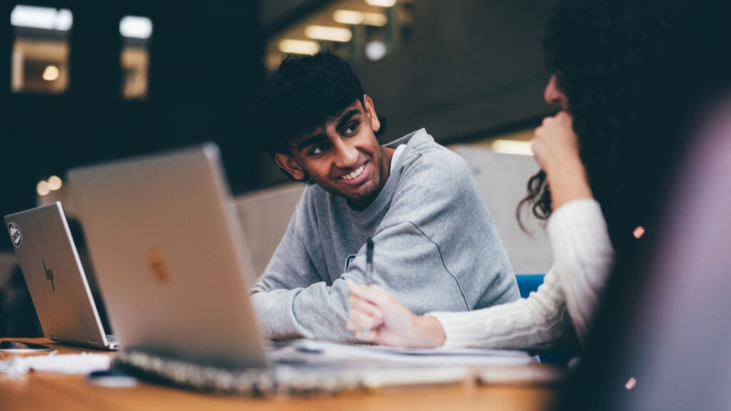 Two students sit side-by-side at a libary table, with open laptops in front of them