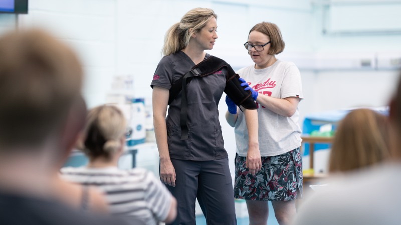 Two women stand at the front of a classroom during a medical training session. One woman, wearing scrubs, demonstrates with her arm in a black medical sling while the other adjusts it. Several seated