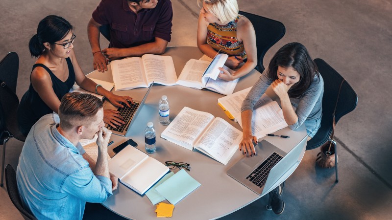 A group of five students sit around a round table, studying together with open books, laptops, notebooks, and water bottles spread out in front of them. They appear to be discussing their work in a c