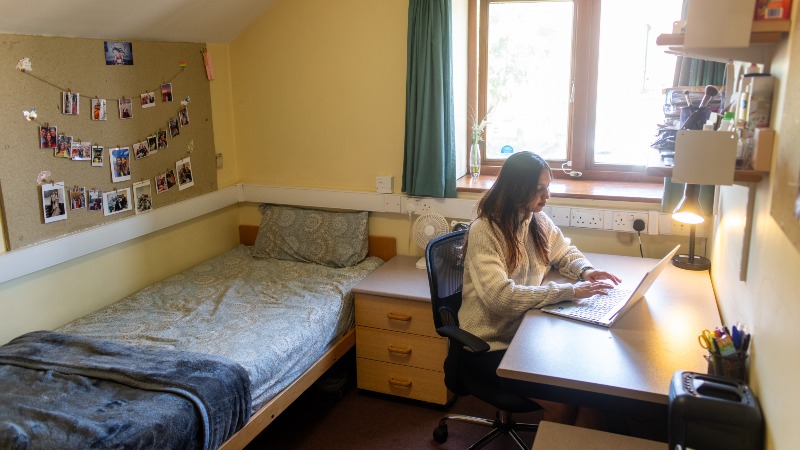 Single bedroom with bed, bedside table and desk. Female student at desk working on laptop.