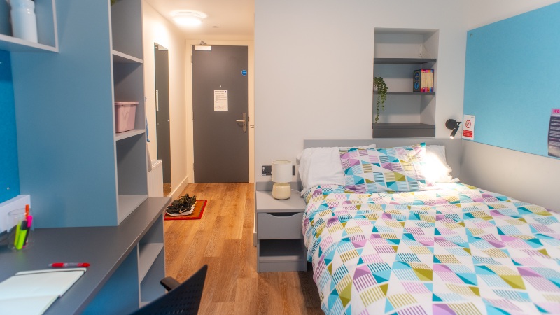 Modern bedroom with light wood flooring. Double bed with multicoloured bedding. Study area, storage and pinboards.