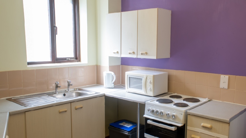 Kitchen corner with bright window, kitchen cabinets, freestanding oven, sink, microwave and kettle.