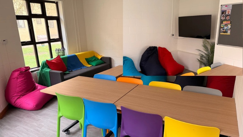 Social space - room with large table and coloured chairs. Sofa and bean bag seating. TV on the wall in the corner. Large windows behind the sofa.