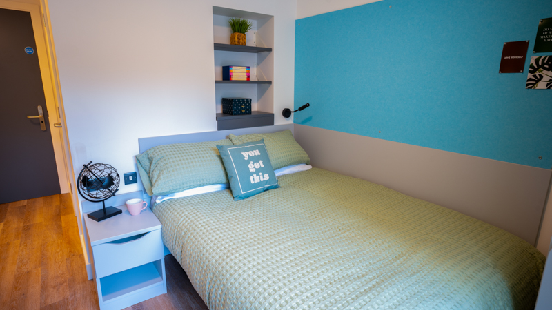 Modern bedroom with light wood flooring. Double bed with blue and white tartan bedding. Green pin board with photos and fairy lights. Modern bed side table and behind the bed shelving.