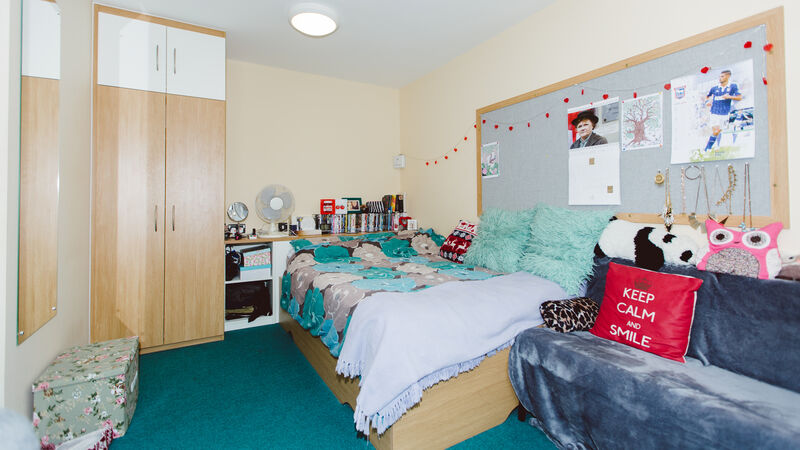 Studio bedroom with double bed, shelving next to the bed, tall wardrobe, pinboard and seating.