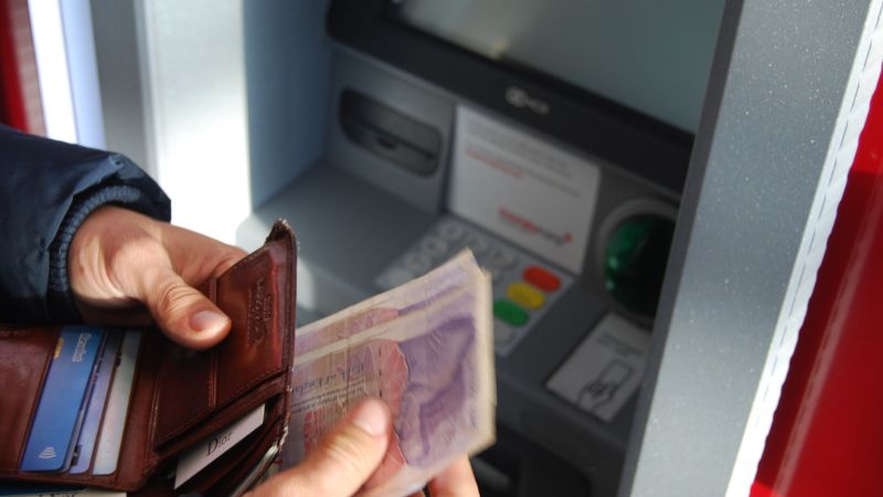 A person's hands holding an open wallet and cash in front of a cash machine