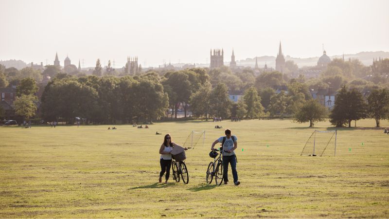 Two students walking with bikes on a sunny day in South Park with a view of the Oxford skyline in the background.