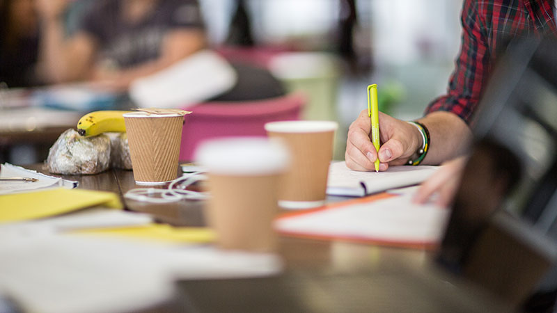 Closeup of a student's hand writing notes with coffee cups in the foreground