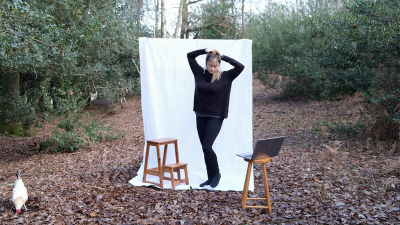 Image of a woman standing in the woods in front of a fabric backdrop, next to a wooden stool with a laptop placed on it