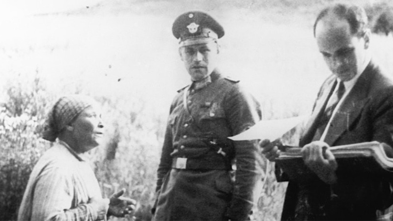 Image of Romani woman standing next to a man in police uniform, and another holding a folder of paperwork 