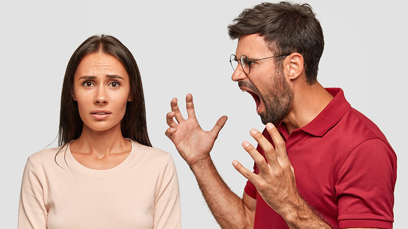Image of a worried looking woman looking into the camera, while a man angrily shouts at her on her left hand side. 
