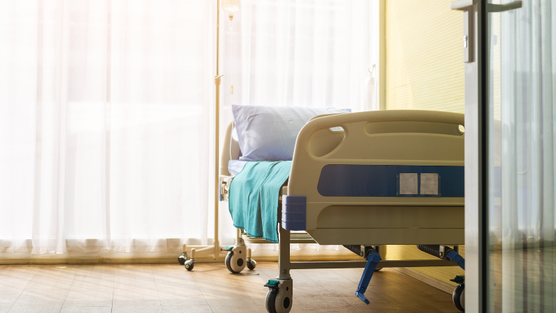 An empty hospital bed in a bright room with sunlight streaming through large windows and thin white curtains.
