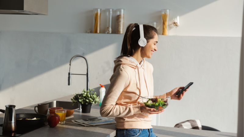 student in a kitchen with headphones on, holding a phone and a bowl of salad